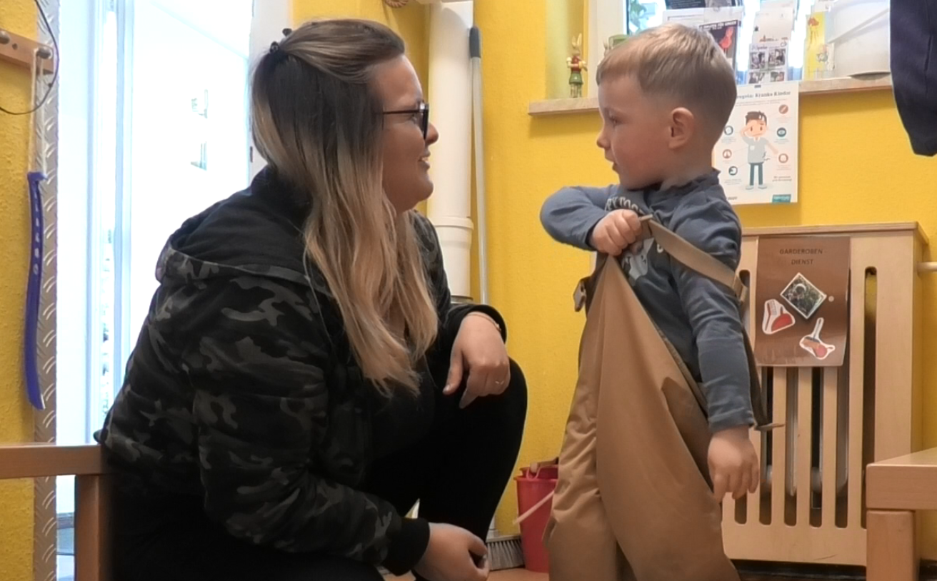 Mate Meo-Element „Positives Leiten“: Für Ludwig (3) ist es gar nicht so einfach, die Regenhose anzuziehen. Erzieherin Raphaela Scherfling bestärkt ihn aufmunternden Gesten und Worten, ohne fordernd oder belehrend zu sein. Seinen Erfolg feiern sie zusammen und er bekommt ein nuanciertes Feedback. Foto: Maria Fleischer